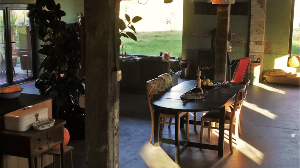 Offener Wohnbereich in einem rustikalen Gebäude mit freiliegenden Mauerwerk, großen Fenstern, die einen Blick ins Grüne bieten, einem schwarzen Esstisch mit Stühlen und Pflanzen als Dekoration. Im Hintergrund befinden sich ein Sofa, ein Holzofen und weitere Details einer gemütlichen Einrichtung.