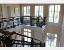 Innenraum eines eleganten Treppenhauses mit cremefarbenen Wänden, großen, sprossenverzierten Fenstern und Balkongeländern aus schwarzem Metall mit Holzhandläufen. Ein großer Strauß Blumen und eine Zimmerpflanze sind dekorativ aufgestellt, während Tageslicht den Raum hell erleuchtet.