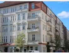 Mehrstöckiges Wohngebäude mit heller Fassade, Balkonen und roten Dachelementen in einer städtischen Umgebung. Vor dem Gebäude stehen Bäume, Straßenlaternen und parkende Autos.