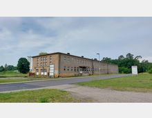 Flaches, rechteckiges, mehrstöckiges Gebäude mit beiger Fassade und zahlreichen Fenstern. Das Gebäude steht in einer offenen, von Grünflächen umgebenen Umgebung, mit einer asphaltierten Straße im Vordergrund und Bäumen im Hintergrund.