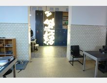 Innenraum eines modern eingerichteten Arbeitsbereichs mit weißen Kachelwänden, Boden aus kleinen Fliesen und schwarzen Spinden im Hintergrund. Zentral befindet sich ein leuchtendes Kunstobjekt mit geometrischen Formen, daneben ein Schreibtisch und Stühle.