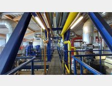 Innenansicht einer technischen Anlage mit Rohrleitungen in verschiedenen Farben (gelb, rot, grau) und blauer Metallstruktur. Die Halle hat eine Decke aus Wellblech, zahlreiche Leitungen und Ventile sowie Beleuchtungselemente.