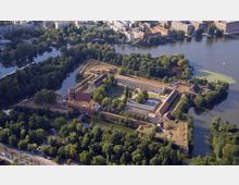 Luftaufnahme einer historischen Festungsanlage, umgeben von Wasser und Bäumen. Die Festung hat eine rechteckige Form mit beeindruckenden Backsteinmauern und Innenhöfen, und ist in eine grüne Parklandschaft eingebettet, mit einer Stadt im Hintergrund.