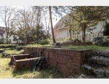 Grüne Gartenlandschaft mit einer niedrigen Backsteinmauer, umrankt von Efeu, und umliegenden Bäumen. Im Hintergrund sind ein Wohnhaus mit schrägem Dach und eine Hecke zu sehen.
