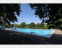 Freibad mit großem rechteckigem Schwimmbecken, umgeben von Bäumen und einer gepflasterten Fläche. Im Hintergrund befinden sich ein Sprungturm, ein Gebäude und grüne Hecken.