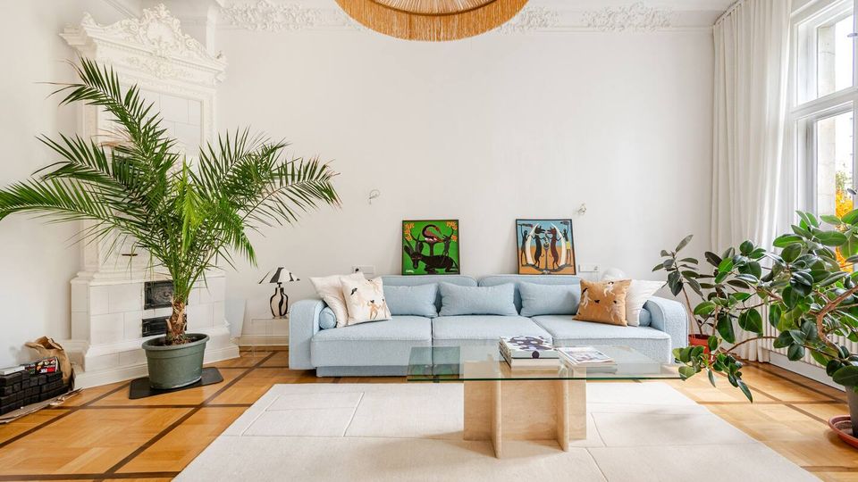 Helles Wohnzimmer mit weißer Stuckdecke, Parkettboden und großem Fenster mit Vorhängen. Vor einer hellblauen Couch stehen ein Glas-Couchtisch und ein heller Teppich; links ist ein weißer Kachelofen, dazu mehrere große Zimmerpflanzen und zwei gerahmte Bilder an der Wand.