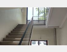 Treppenhaus mit einfacher Betonstiege und Metallgeländern, das von einem großen Fenster mit vertikalen Sprossen und Sicht auf Bäume und geparkte Autos beleuchtet wird. Die Wände sind in hellem Beige gestrichen, und ein Heizkörper ist unter dem Fenster sichtbar.