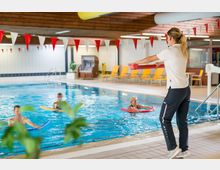 Eine Schwimmhalle mit einem rechteckigen Becken, in dem drei Personen mit Schwimmnudeln Übungen machen, während eine Trainerin am Beckenrand Anweisungen gibt. Im Hintergrund sind gelbe Liegestühle, ein Strandkorb und geflieste Wände zu sehen.