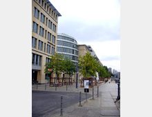 Straßenansicht eines modernen Büro- und Geschäftsviertels mit einem Gebäude aus Glas und Steinfassade auf der linken Seite. Entlang der Straße stehen junge Bäume, Fahrradständer und Poller, dahinter sind Sitzbereiche mit weißen Tischen und Stühlen sichtbar.