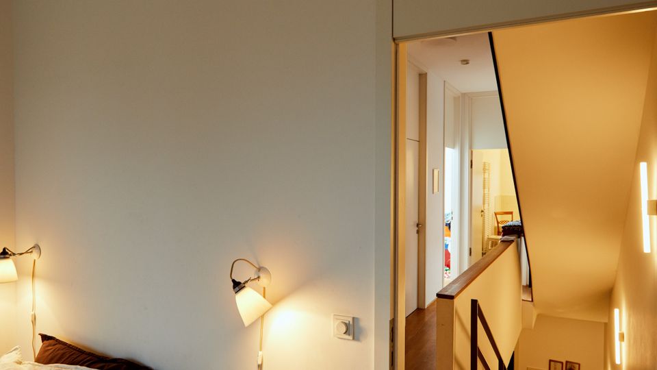 A modern interior showing a small bedroom with a white wall, a bed with neutral bedding, and wall-mounted reading lights. Adjacent to the bedroom is a hallway with wooden floors leading to a staircase with a black metal railing, illuminated by warm wall sconces.