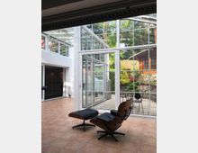 Moderne Inneneinrichtung mit einem Sitzbereich bestehend aus einem Lounge-Sessel und Hocker auf einem Terrakotta-Fliesenboden. Der Raum verfügt über große Glaswände, die eine Terrasse mit Gartenmöbeln und üppiger Bepflanzung sowie umliegende Gebäude und Bäume sichtbar machen.