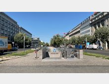 Eingang zur U-Bahn-Station "Unter den Linden" in Berlin, umgeben von großen Bürogebäuden und Bäumen entlang der Straße. Im Vordergrund befinden sich Rolltreppen und ein Fahrstuhl, während der Bereich von befahrbaren Straßen und parkenden Fahrzeugen eingerahmt wird.