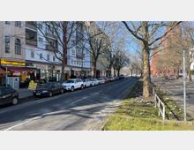 Straßenansicht in einer Stadt mit parkenden Autos entlang des Gehwegs, Wohngebäuden mit Geschäften im Erdgeschoss auf der linken Seite und einer Baumreihe entlang der Straße. Im Vordergrund blühen Krokusse auf einer Grünfläche, und im Hintergrund ist eine rote Backsteinkirche sichtbar.