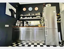 Moderne Küche mit dunklen Wänden, einer Arbeitsfläche aus Holz, Edelstahlfronten und einem großen silbernen Kühlschrank. Darüber befinden sich offene Regale mit Geschirr und Dekoration, die schwarz-weißen Fliesen ergänzen den stilvollen Look.