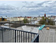 Dachterrasse mit Holzboden und schwarzen Metallgeländern, ausgestattet mit weißen und türkisfarbenen Sitzmöbeln. Im Hintergrund sind Häuser mit roten Ziegeldächern und eine Backsteinkirche mit grünem Turm vor einem bewölkten Himmel zu sehen.