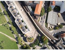 Luftaufnahme einer städtischen Umgebung mit einem Bahnsteig und einer erhöhten Bahntrasse, die von einer gelben U-Bahn passiert wird. Umgeben sind die Schienen von modernen Wohngebäuden, einem parkähnlichen Grünbereich sowie weiteren Gebäuden und Straßen.