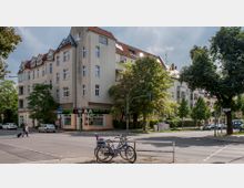 Wohngebäude an einer Straßenecke mit mehreren Etagen, sandfarbener Fassade und roten Ziegeldächern, darunter ein Geschäft mit Beschriftung „Pizza Pasta Burger“. Im Vordergrund stehen zwei Fahrräder an einem Geländer, Bäume säumen die Straße und es sind parkende sowie fahrende Autos sichtbar.