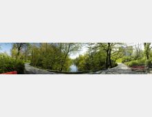 Panorama eines Parks mit einem schmalen Weg, der sich um einen kleinen, von Bäumen umgebenen Teich windet. Auf beiden Seiten des Weges stehen rote Parkbänke, und im Hintergrund ist ein modernes Gebäude zwischen den Bäumen sichtbar.