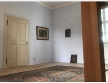 Ein schlichtes Zimmer mit weißen Wänden, einer Holztür und einem Fenster mit hellem Licht. Auf dem Boden liegt ein großer gemusterter Teppich, und an der Wand hängen Bilder sowie eines, das angelehnt am Boden steht.