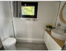 Modernes Badezimmer mit weißer Toilette links, einem schmalen Fenster mit grauem Vorhang in der Mitte und einem Waschbecken auf einer Holzplatte rechts, daneben ein Blumenstrauß in einer Vase. Der Boden ist mit grauen Fliesen ausgelegt, und die Wände sind weiß.