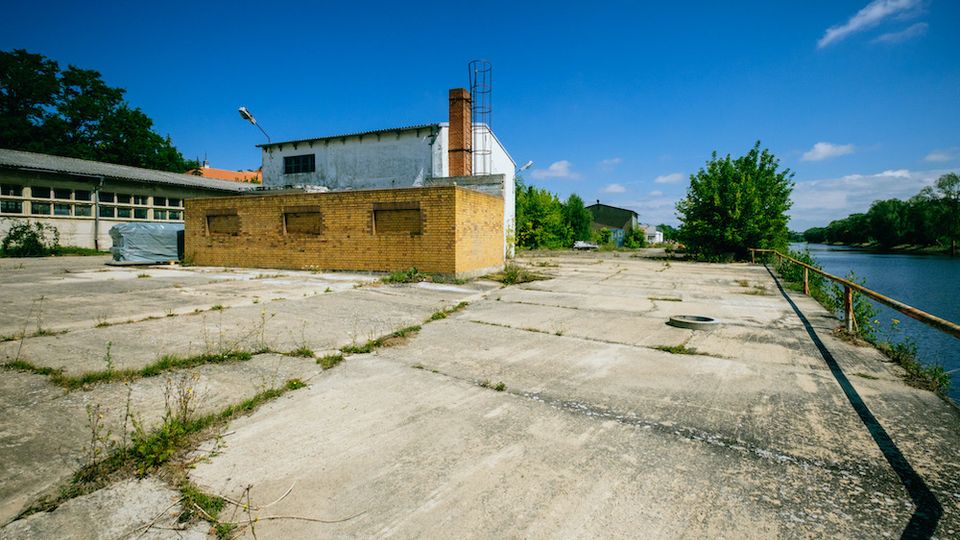 Industriegelände mit leerer Betonfläche, einem kleinen Backsteingebäude und einem größeren weißen Gebäude im Hintergrund; rechts verläuft ein Fluss mit Geländer entlang der Uferseite, umgeben von Bäumen und Vegetation.