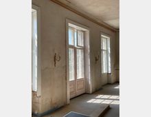 Ein heller Raum mit hohen, verzierten Doppelfenstern und einer Tür mit Glaspanels sowie hölzernen Details. Die Wände sind schlicht, leicht beschädigt, und an beiden Seiten der Fenster hängen dekorative Wandkerzenhalter.