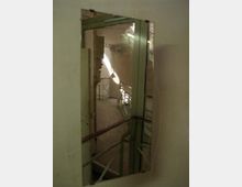 Ein Spiegel an einer Wand reflektiert einen Teil eines Gebäudeflurs mit metallenen Geländern, einer Glasabtrennung und einem Fenster, durch das Tageslicht einfällt. Die Umgebung wirkt industriell, mit sichtbaren Rohren und kahlen Wänden.