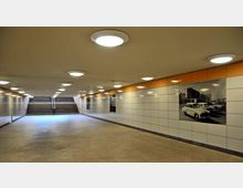 U-Bahn-Unterführung mit beige gefliesten Wänden, beleuchtet von runden Deckenlampen. An den Wänden befinden sich historische Schwarz-Weiß-Bilder, und im Hintergrund führen Treppen ins Freie.