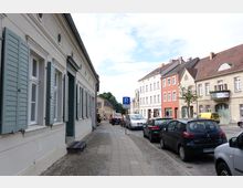 Straßenszene in einer Kleinstadt mit Kopfsteinpflaster und Gehweg; links ein Gebäude mit hellen Wänden und grünen Fensterläden, rechts eine Reihe von mehrstöckigen Wohnhäusern in verschiedenen Farben. Geparkte Autos stehen entlang der Straße, und ein Verkehrsschild für Parken ist sichtbar.