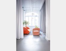 Heller, moderner Loungebereich mit großer bogenförmiger Fensterfront, grauen Vorhängen und glattem Boden. In der Mitte stehen ein orangefarbenes Sofa und ein rosafarbener Sessel auf einem gemusterten Teppich, ergänzt durch Hängelampen und Zimmerpflanzen.