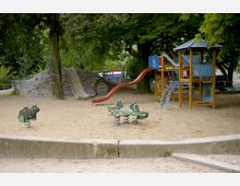 Ein Spielplatz mit einer Sandfläche, umgeben von Bäumen. Zu sehen sind eine Wippe, eine wellenförmige Rutsche, ein Klettergerät mit Rutsche und ein gemauerter Hügel mit integrierter Rutsche und Tunnel.