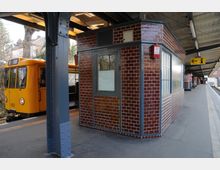 Bahnhof mit gelber U-Bahn, die an einem Bahnsteig hält. Im Vordergrund ein kleines Gebäude mit glänzenden, braunen Fliesen, darüber ein überdachtes Dach mit Metallstützen.