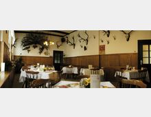 Ein traditioneller Speiseraum mit Holztäfelung und weißen Tischdecken, dekoriert mit Wandtrophäen wie Hirschgeweihen und einem präparierten Pfauenfächer. Holzmöbel und eine einfache Beleuchtung prägen das Ambiente.