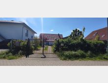 Einfamilienhaus mit rotem Dach hinter einem Metalltor, umgeben von Garten und Büschen. Links befindet sich ein helles Gebäude, rechts ein weiteres Haus mit rotem Dach, und der Himmel ist klar mit wenigen Wolken.