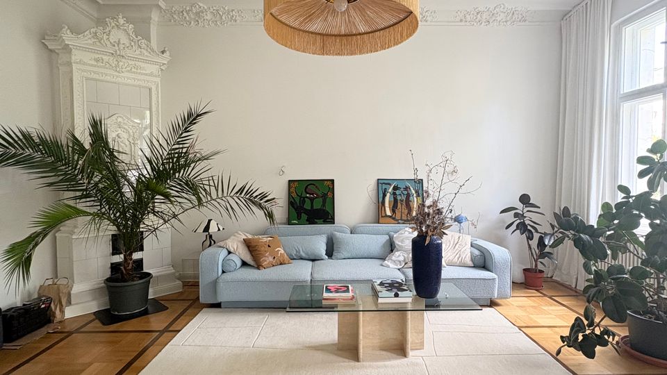 Helles Wohnzimmer mit hohen Decken, Stuckverzierungen und großem Deckenrosetten-Leuchtenelement, mit Parkettboden und großem hellen Teppich. Vor einer hellblauen Sofa-Sitzgruppe steht ein niedriger Glastisch; große Zimmerpflanzen und bodentiefe Fenster mit weißen Vorhängen rahmen den Raum.
