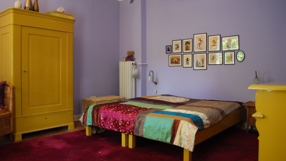 Schlafzimmer mit lavendelfarbenen Wänden, rotem Teppichboden und einem Bett mit bunter Patchworkdecke. Links steht ein großer gelber Kleiderschrank, an der Wand hängen mehrere gerahmte Bilder, daneben sind ein Heizkörper und ein kleiner Nachttisch zu sehen.