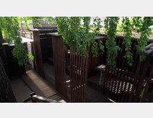 Holzterrasse mit hohen, vertikalen Holzlattenstrukturen, die wie Abtrennungen wirken, teilweise von hängendem Grün bedeckt; im Hintergrund weitere Holz- und Pflanzenstrukturen.