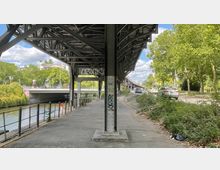 Überdachter Gehweg unter einer Stahlbrücke mit verschraubten Metallträgern und Graffiti, links ein Geländer entlang eines Kanals, rechts eine befahrene Straße mit Autos und Bäumen im Hintergrund.