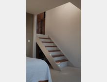 Eine einfache, schmale Holztreppe mit weißen Seitenwangen und dunkelbraunen Trittstufen führt an einer cremefarbenen Wand entlang nach oben. Im Vordergrund ist eine Ecke eines Bettes mit weißer Bettdecke zu sehen.