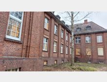 Backsteingebäude mit mehreren rechteckigen Fenstern, von denen einige mit Holzplatten verschlossen sind. Vor dem Gebäude steht ein kahler Baum, während im Vordergrund Gras und Büsche zu sehen sind.
