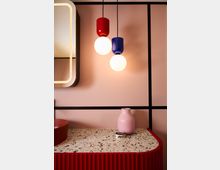 Eine moderne Innenraumgestaltung mit einer Tischfläche aus Terrazzo, einer roten geriffelten Front, einer rosafarbenen Vase und einer kleinen Dose mit der Aufschrift "IMMER". Darüber hängen zwei runde Hängeleuchten mit bunten Fassungen in Rot und Blau vor einer beigen Wand mit schwarzen Linien.