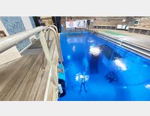 Ein Innenraum mit einem großen, rechteckigen Tauchbecken mit tiefblauem Wasser, umgeben von einem hölzernen Laufsteg und metallenen Geländern. Im Hintergrund sind Tauchausrüstung, Pressluftflaschen und Werbebanner sichtbar.