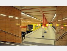 U-Bahn-Station mit orangefarbenen Wandfliesen, gelben Informationsschildern und einem gelben U-Bahn-Zug der Linie U5 am Bahnsteig. Die Decke ist mit parallelen Lichtbändern beleuchtet, und rote Pfeiler teilen die Bereiche.
