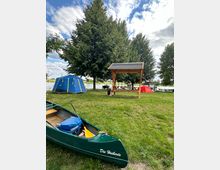 Ein Campingplatz mit grüner Wiese, auf der mehrere Zelte stehen. Im Vordergrund ein grünes Kanu mit der Aufschrift „Die Heiberts“, daneben ein hölzerner Unterstand und ein Picknicktisch; im Hintergrund Bäume und ein Gewässer.