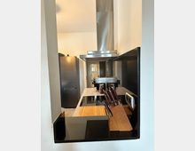 Moderne Küche mit offenem Durchgang, einem Herd mit schwarzer Glaskeramikoberfläche und Edelstahldunstabzugshaube. Im Vordergrund steht ein Messerblock, im Hintergrund sind ein Kühlschrank und eine Kaffeemaschine sichtbar.