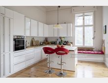 Helle Küche mit weißen Einbau-Schränken und Arbeitsplatten aus Holz, zentralem Küchenblock mit zwei roten Barhockern, großem Fenster mit Blick ins Freie und einer integrierten Sitzbank am Fenster. Der Raum hat Holzdielenboden und ist schlicht und modern gestaltet.