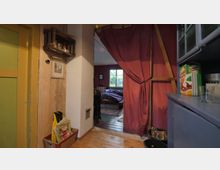 Ein kleiner Innenraum mit Holzboden, einer gelben Tür auf der linken Seite und einem roten Vorhang, der einen Übergang zu einem Schlafzimmer mit Fenster und Bett freigibt. Rechts steht ein blaues Regal mit Lebensmitteln, darunter eine Packung Schokocerealien.