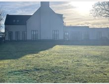 Moderne Gebäude mit weißer Fassade und großen, bodentiefen Fenstern, umgeben von einer Rasenfläche. Im Hintergrund sind Bäume und ein heller, sonniger Himmel zu sehen.