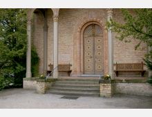 Eingang einer historischen Kirche mit einer Doppeltür aus Holz, verziert mit kreisförmigen Ornamenten. Vor der Tür befinden sich eine kleine Treppe, Zierpflanzen in Blumentöpfen und zwei Sitzbänke, eingerahmt von Säulen aus Stein.