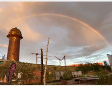 Ein hoher, runder Backsteinturm mit spitzem Dach steht auf einer leicht bewachsenen Anhöhe, flankiert von mehreren Baukränen im Hintergrund. Über die Szene spannt sich ein Regenbogen vor wolkigem Himmel, während im Vordergrund ein Gartenbereich mit Pflanzen und diversen Objekten zu sehen ist.